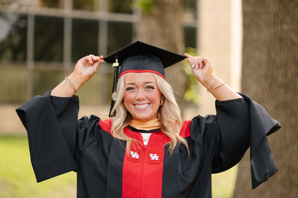 Smiling graduate in black cap and gown with red trim adjusts tassel outdoors during graduation photo session.