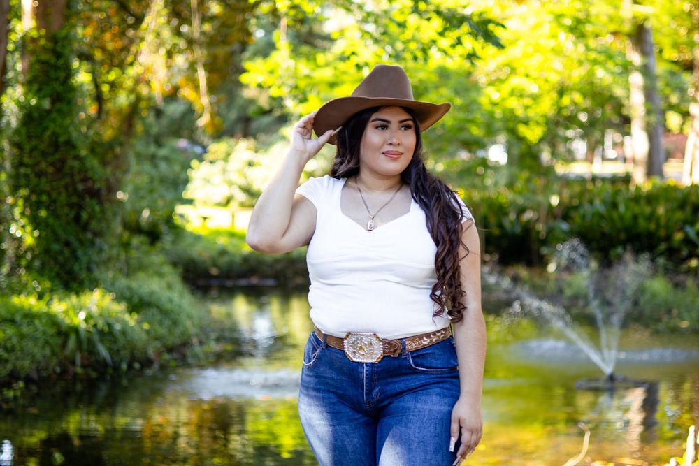 Woman in white top, jeans, and brown cowboy hat posing near a pond in a lush green park.