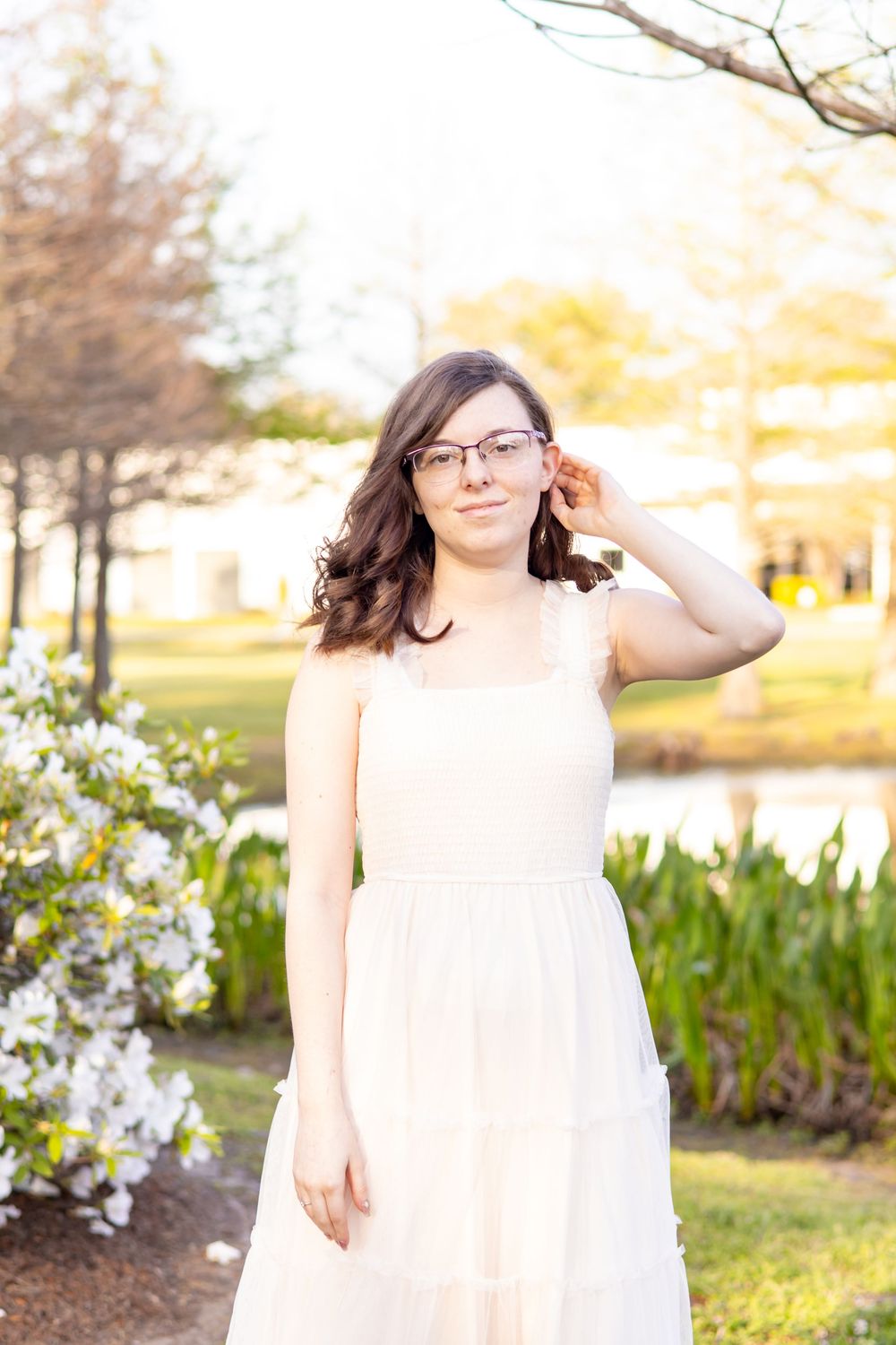 Young woman in white dress adjusting glasses outdoors near blooming flowers on a sunny spring day.