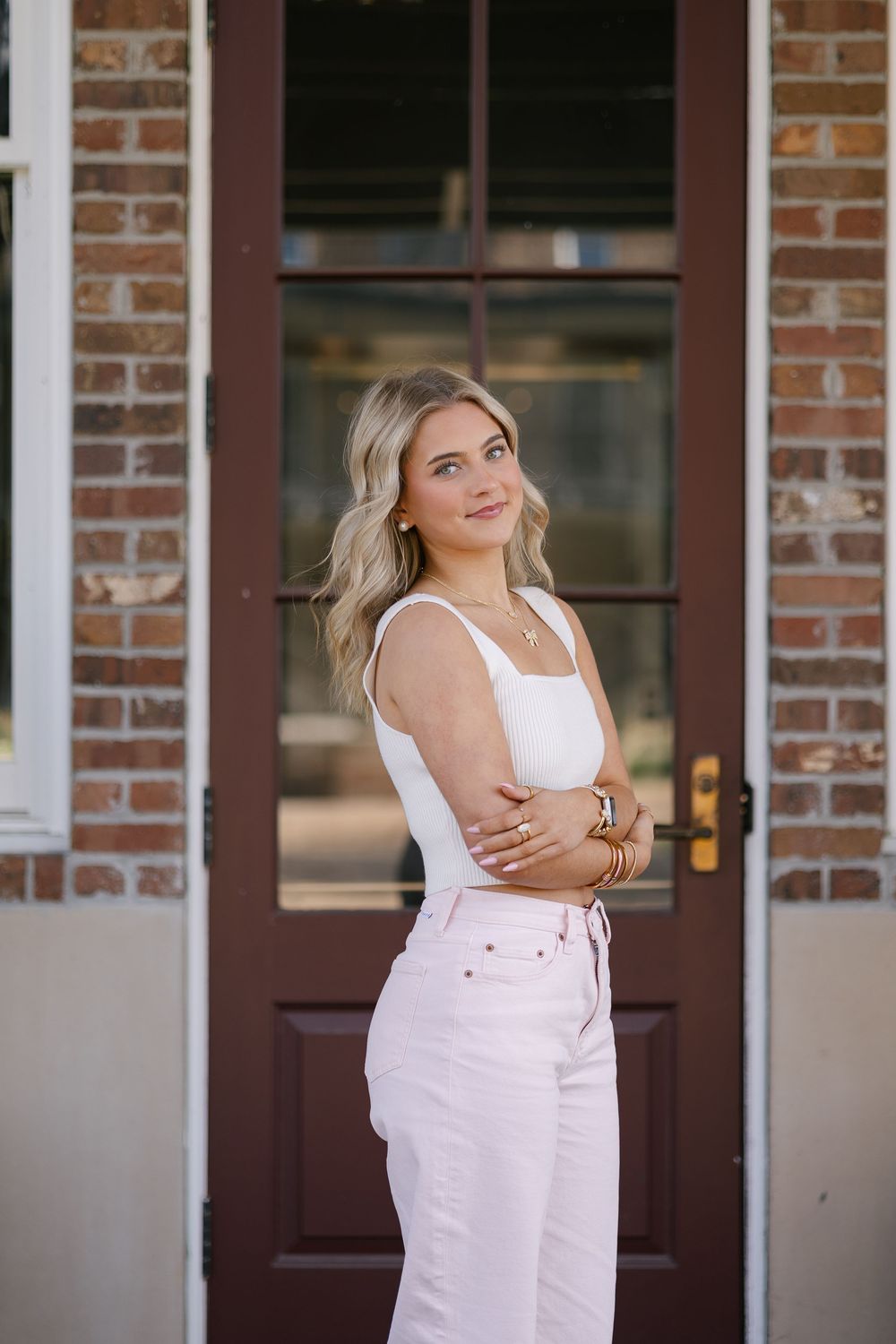 A person in a white tank top and light colored pants poses in front of a brick building with a dark wooden door.