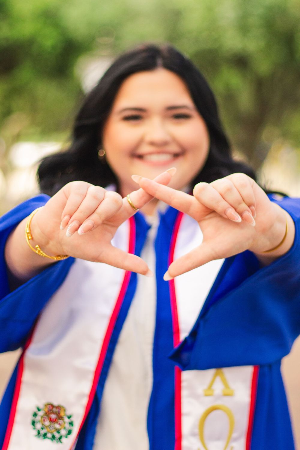 Graduate in blue academic regalia wearing cultural stoles makes CHI-O gesture with hands.