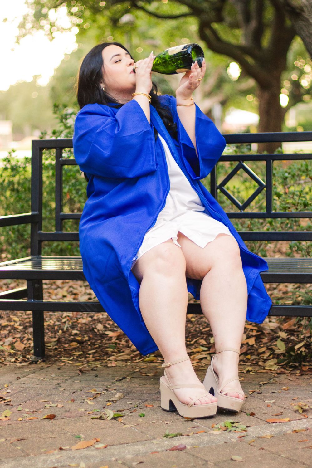 Person in bright blue coat and beige heels sits on park bench drinking from green glass bottle in outdoor setting.