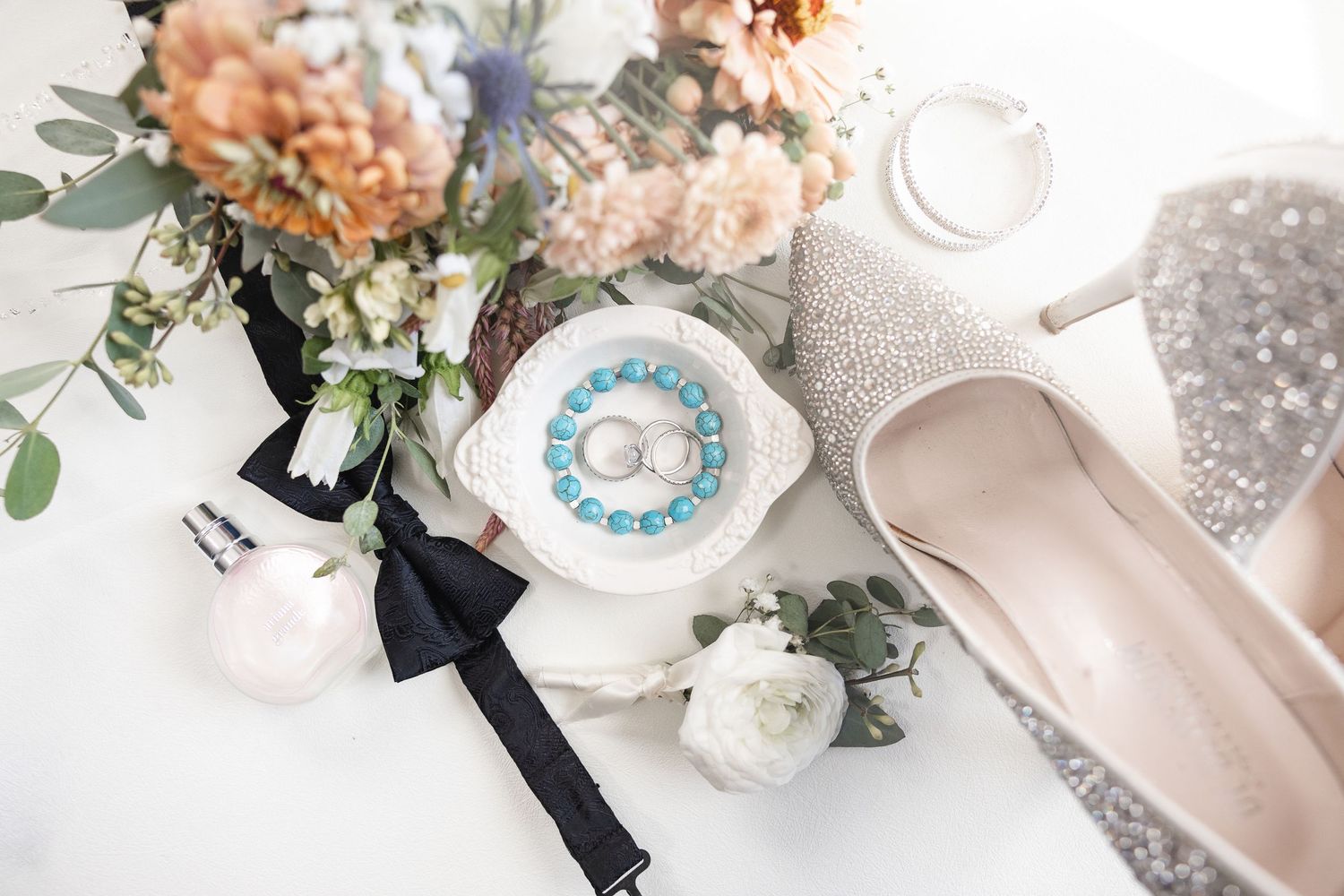 Wedding flat lay with floral bouquet, glittery heels, ring dish, bow tie, and pearl accessories on white background.