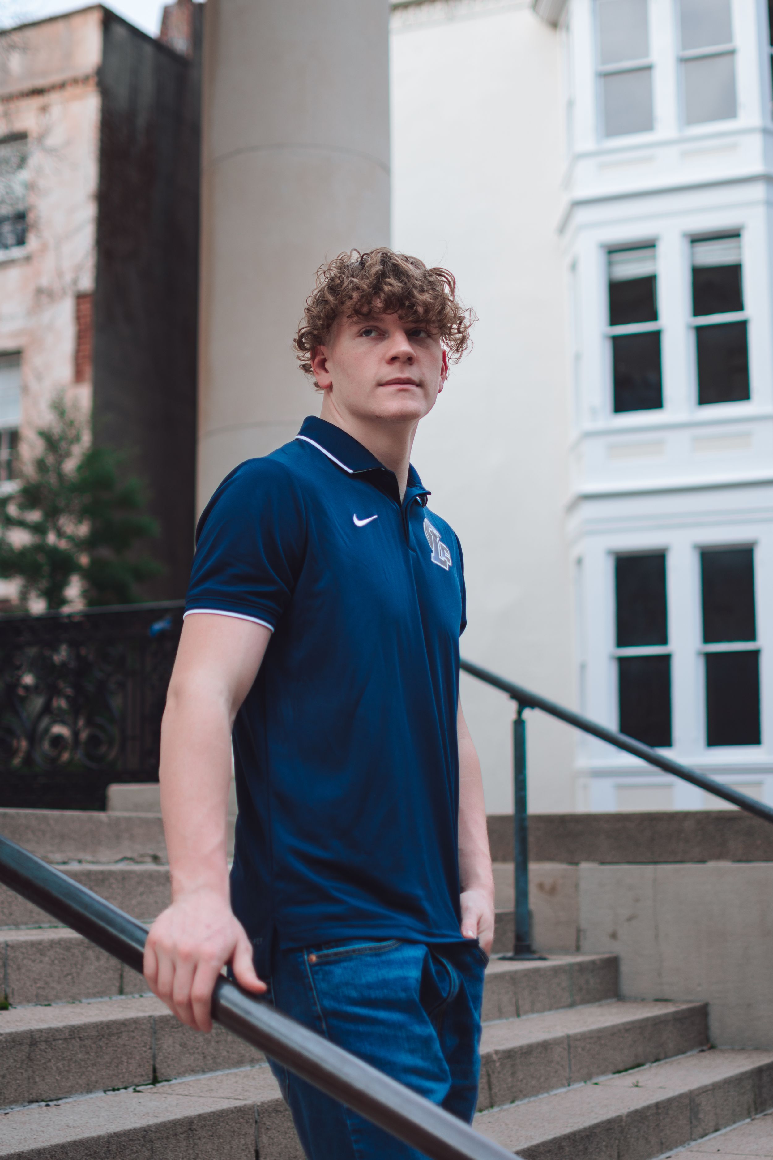 A person with curly hair wearing a navy Nike polo shirt stands on stone steps beside a white building.