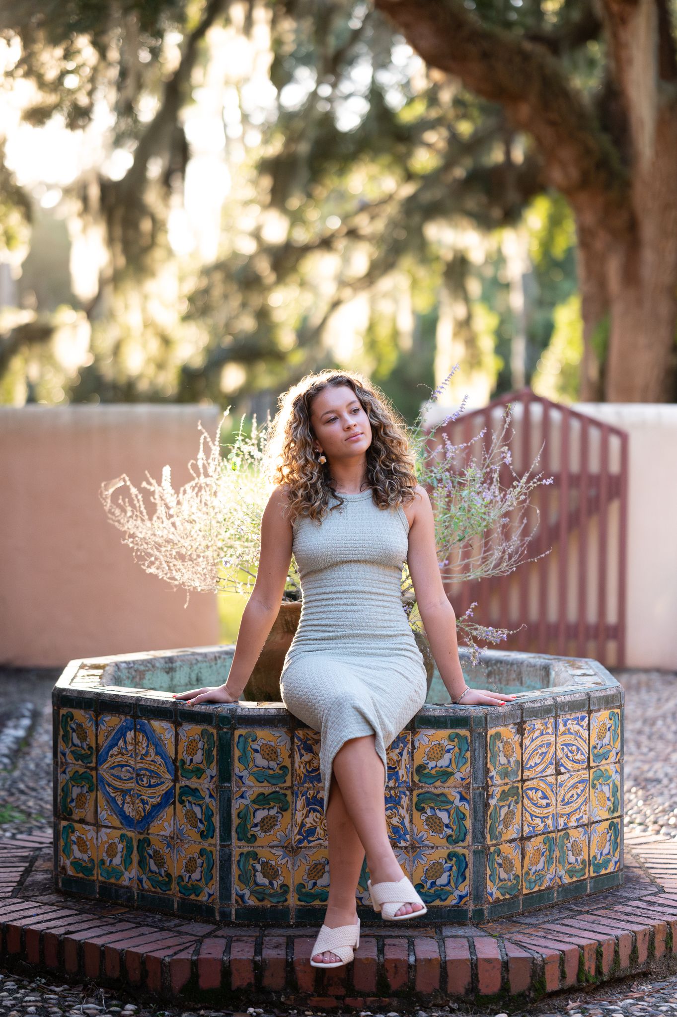 A person in grey dress sits on decorative tiled fountain edge in garden setting.
