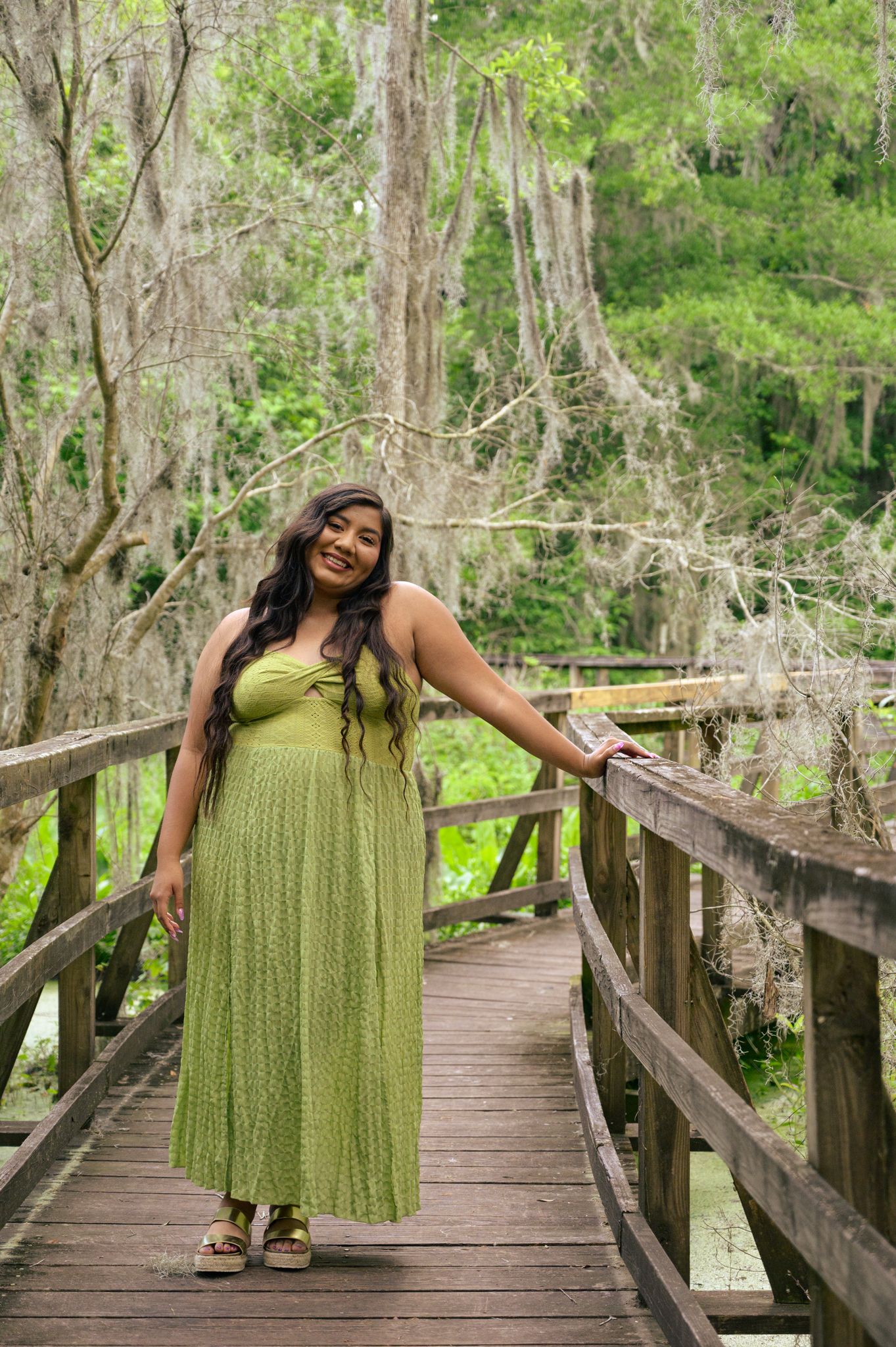 Woman in lime green maxi dress poses on wooden boardwalk through forest.