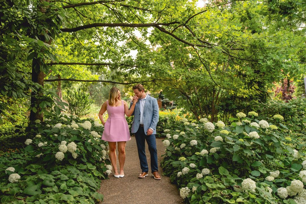Couple walking hand in hand through lush garden path lined with white hydrangeas and green trees.