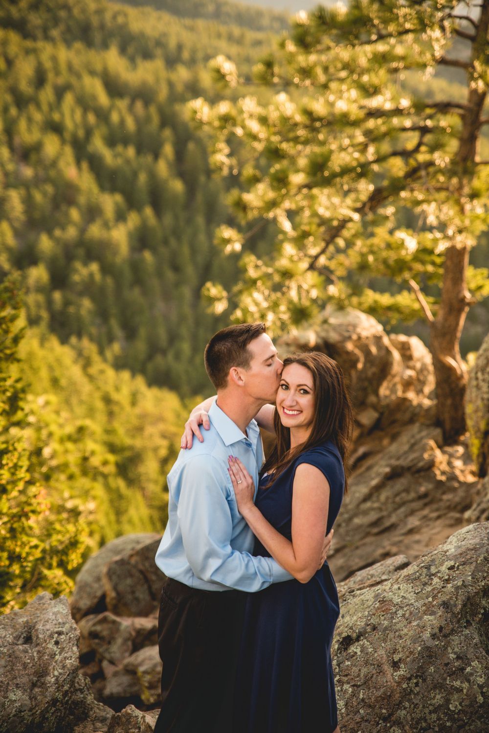 Couple embraces on mountain overlook during golden hour with forest valley below.