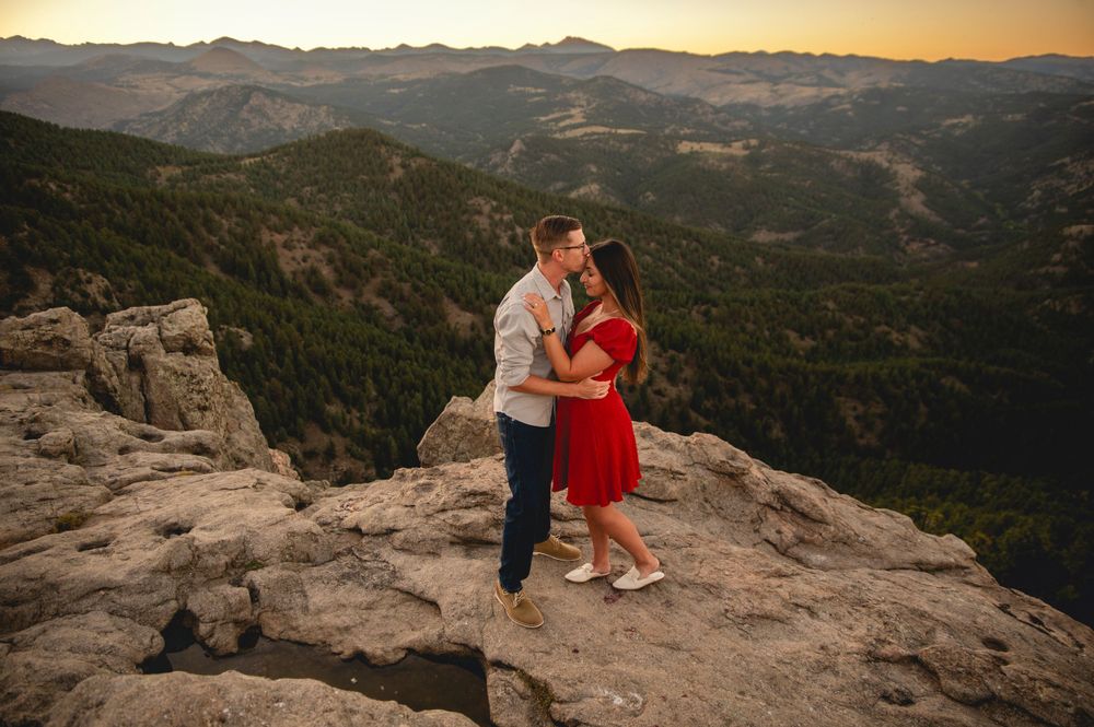 A romantic sunset engagement photo on a rocky mountain overlook with dramatic mountain ranges in the background.