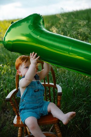 Baby in denim overalls plays with green slide while sitting in wooden chair outdoors.