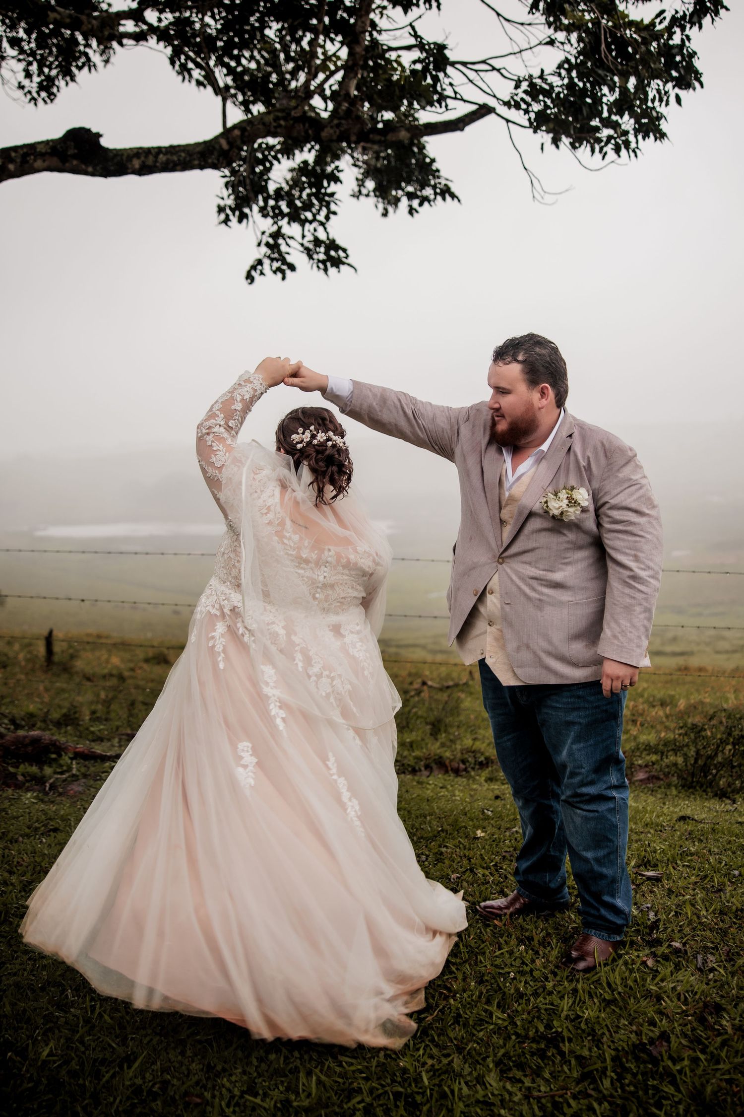 A couple dances together under a tree branch on their wedding day in the foggy countryside.