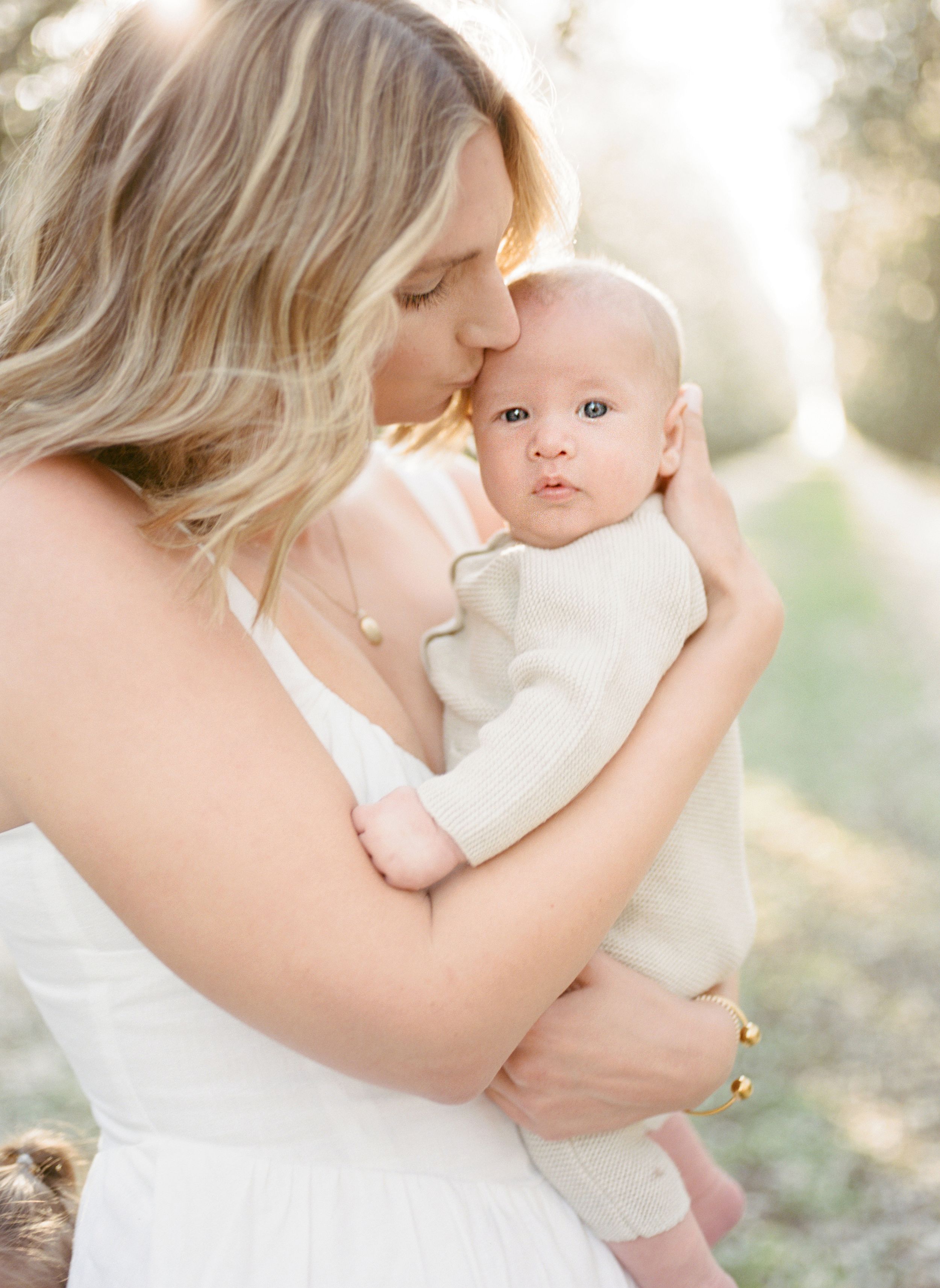A tender moment captured in soft sunlight as a baby in white is held and kissed in a gentle embrace.By Chelsee Rawe Photography.