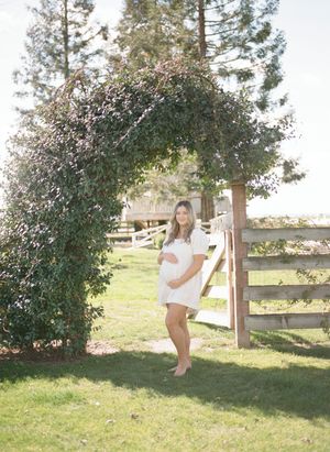 A pregnant woman in a white dress stands beneath a tree arch next to a rustic wooden fence in a sunny garden setting.Film by Chelsee Rawe