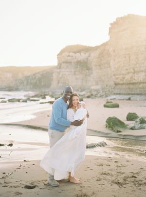 A couple embraces on a scenic beach with dramatic coastal cliffs during golden hour at sunset film by Chelsee Rawe Photography..