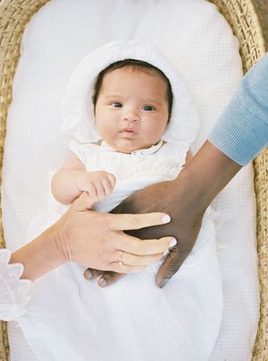 Baby wearing white outfit lies in wicker bassinet while adult hands gently touch their side film by Chelsee Rawe Photography..