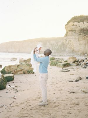 A person in a light blue shirt holds a baby against a backdrop of coastal cliffs and beach at sunset film by Chelsee Rawe Photography..