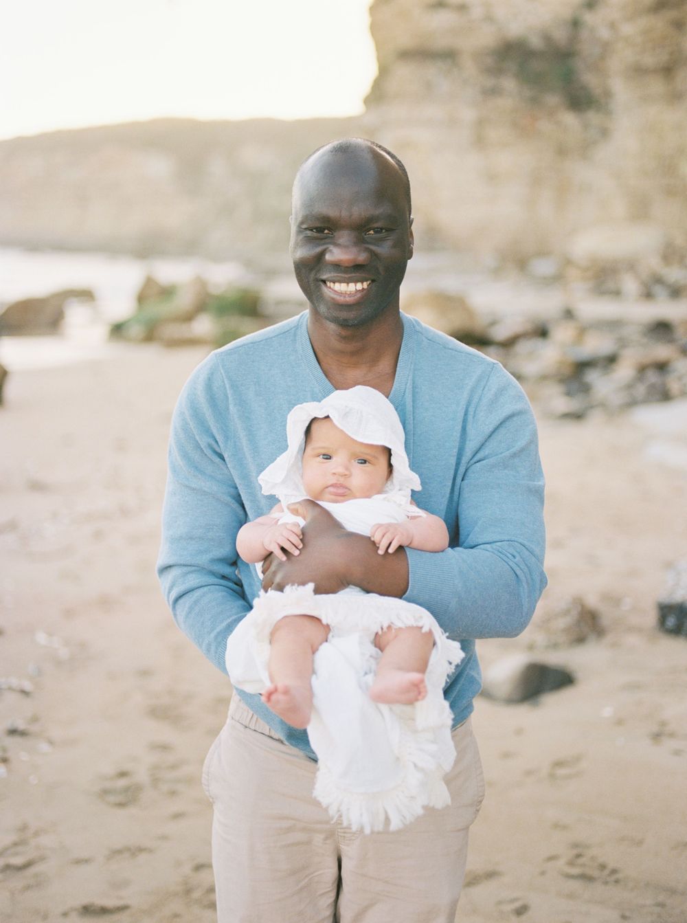 A person in a blue sweater holds a smiling baby in white clothing on a beach with rocky cliffs in the background film by Chelsee Rawe Photo.