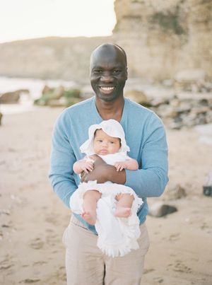A person in a blue sweater holds a smiling baby in white clothing on a beach with rocky cliffs in the background film by Chelsee Rawe Photo.