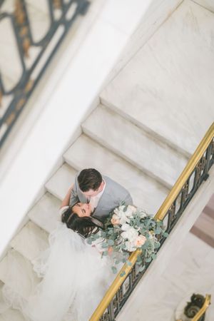 Bride and groom share intimate moment on white marble staircase with eucalyptus bouquet and ornate gold railing.By Chelsee Rawe Photography.