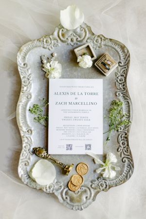 Wedding invitation displayed on ornate white vintage tray with white flowers, gold coins and floral accents as styling details.