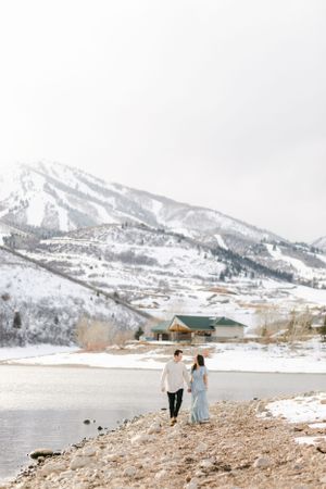 A snowy peak in the looming background with a couple walking along a snowy lake's edge for an engagement session by Chelsee Rawe