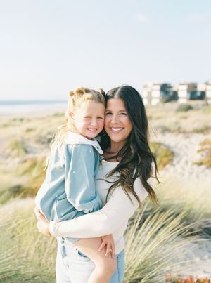 Smiling woman with long dark hair holding a young blonde girl in a denim jacket on a sunny beach with dune grass and coastal buildings.