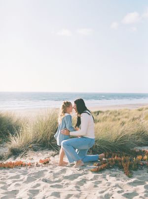 Mother and young daughter share a tender moment on a sandy beach with tall dune grass, with the ocean and soft blue sky behind them.