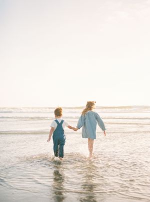 Two young children hold hands while wading through shallow ocean water on a sunny beach at golden hour.