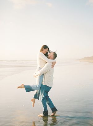 Barefoot couple in jeans and light shirts embracing as he lifts her on a sunlit beach, with gentle waves and soft golden light.