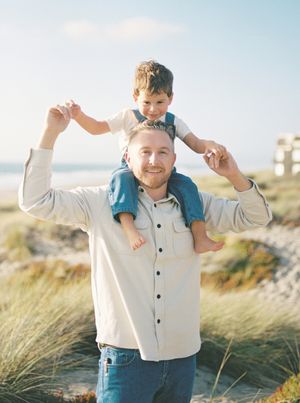 Smiling man in a cream button-up shirt carries a laughing young boy on his shoulders on a sunny beach with dune grass.
