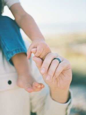 Adult hand with a dark ring gently holding a toddler's tiny hand and bare foot against a soft, blurred beach background.