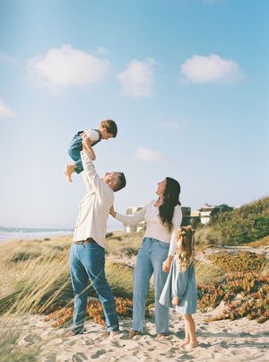 Family of four in light denim and white outfits plays on a sandy beach as a dad lifts a laughing toddler into the air under a blue sky.