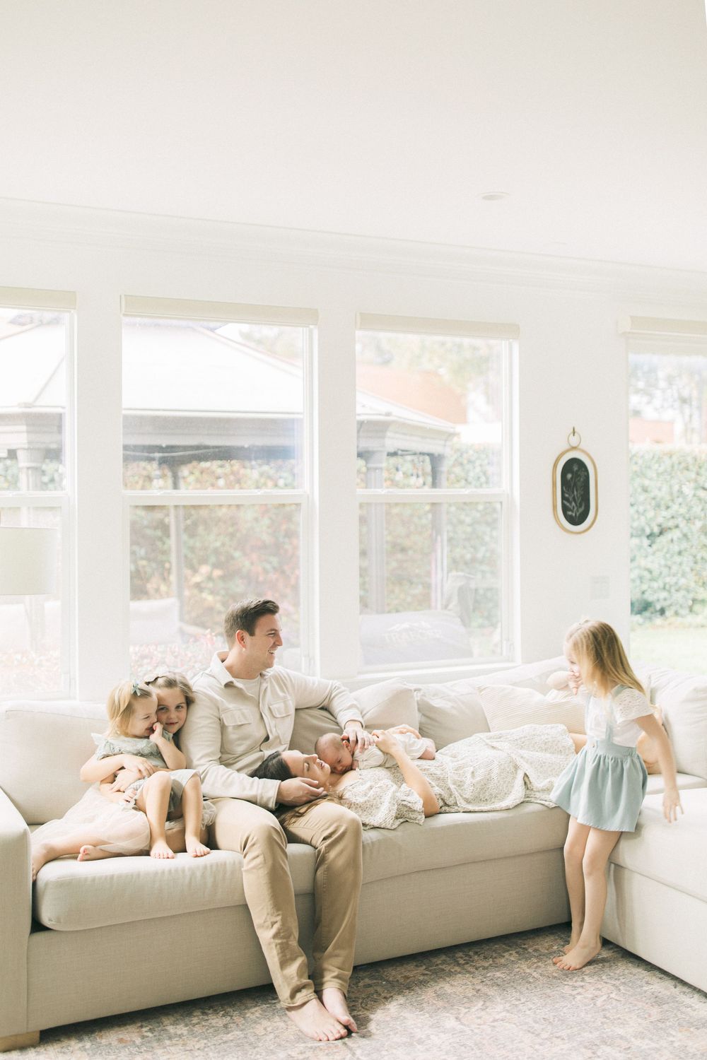 A family lounges together on a long white couch in a bright sunlit room with large windows.