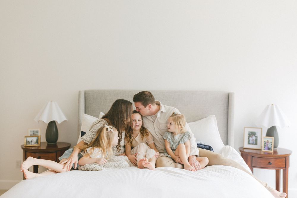 A series of intimate family photos showing parents and children lying together on a white bed in casual poses.