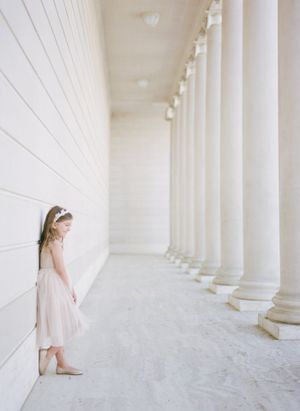 Young girl in a light pink dress stands contemplatively in a white columned hallway with classic architecture.By Chelsee Rawe Photography.
