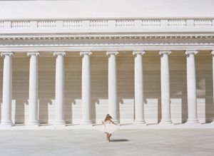 Distant silhouette twirls in a light dress amidst grand white classical columns of a majestic colonnade.By Chelsee Rawe Photography.