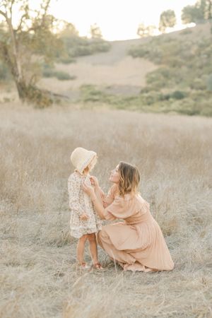 A tender moment captured in a golden field as a figure in peach dress kneels beside small child in floral dress at sunset.By Chelsee Rawe Ph