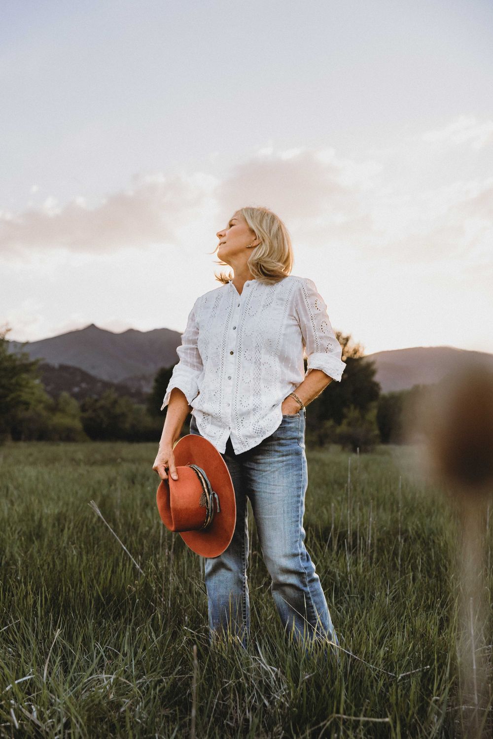 A woman in a white button-down shirt and jeans stands in a grassy field at sunset with mountains in the background.