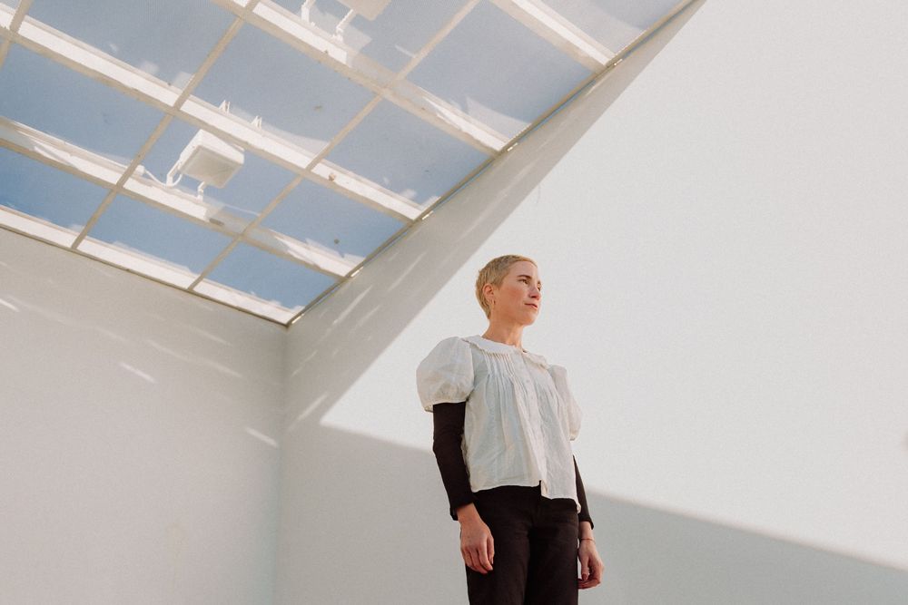 Person in white shirt and black pants stands in sunlit room with skylights casting dramatic shadows on white walls.