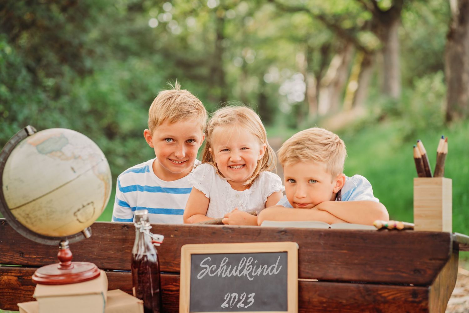 Three students smiling at vintage desk with globe and chalkboard in forest setting.