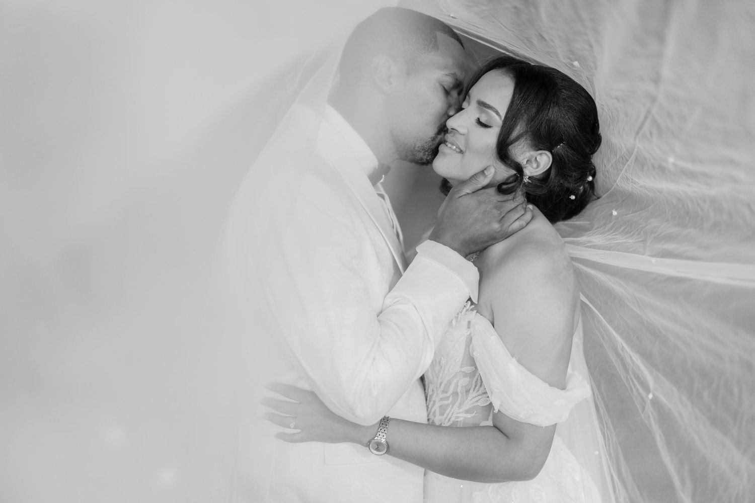 Black and white photo of groom tenderly kissing bride beneath a veil.