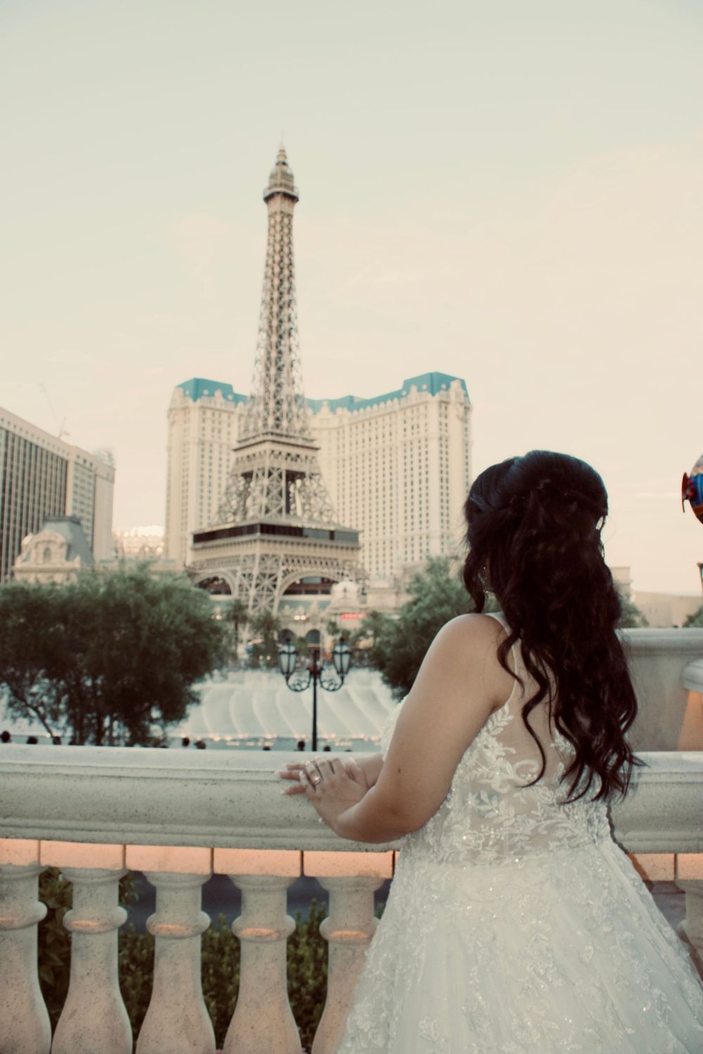 Bride in white lace wedding gown gazes at the Paris Las Vegas Eiffel Tower replica from a balcony.