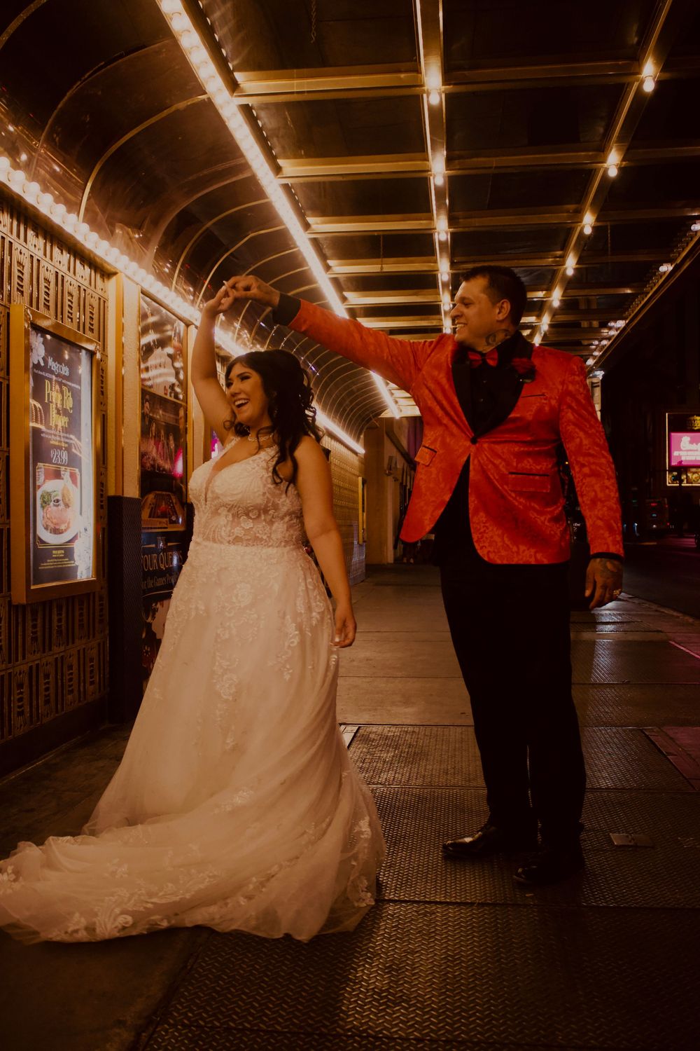 Bride in white gown twirls under groom's hand in red sequin tuxedo outside a lit theater marquee at night.
