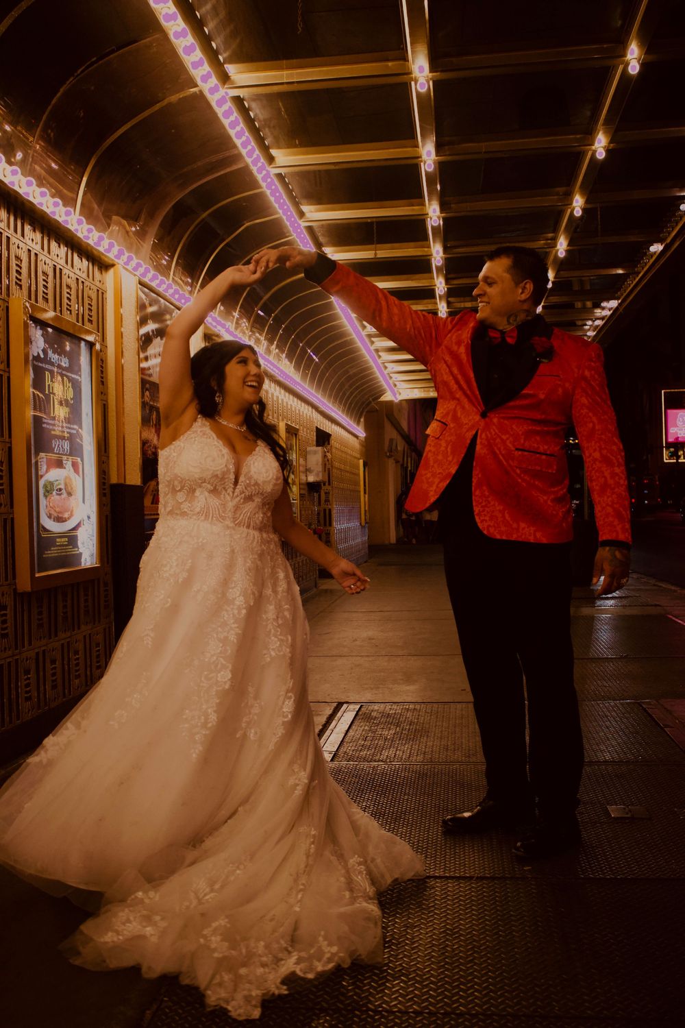 Bride in white lace gown and groom in red blazer dancing under glowing arcade lights.