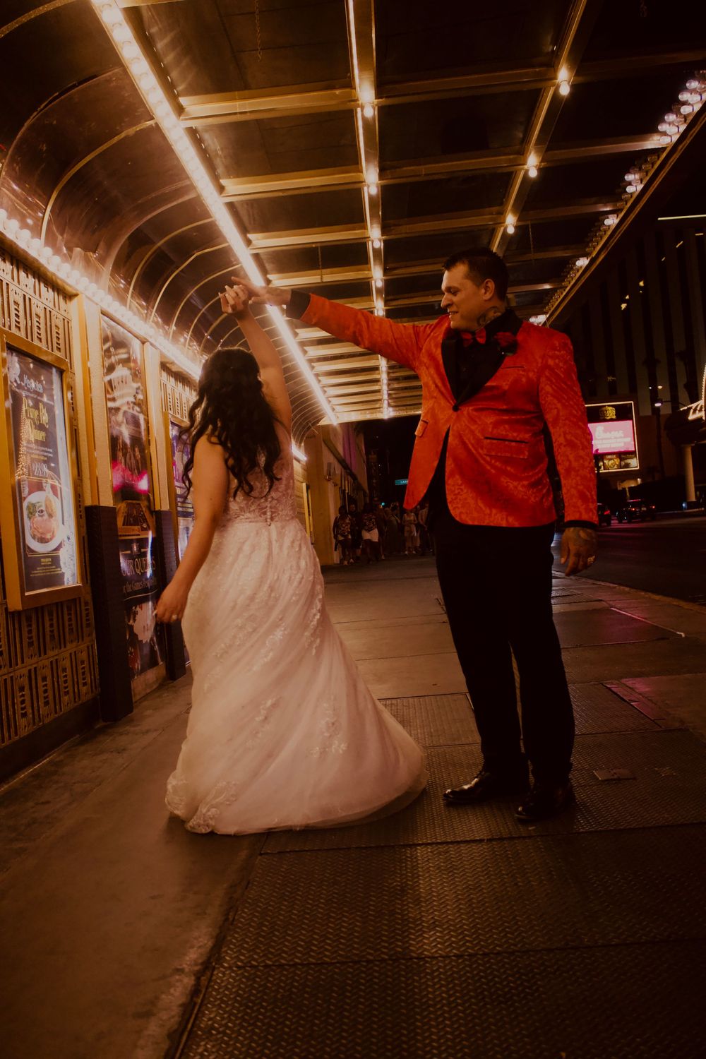 Groom in red sequin blazer twirling bride in white lace wedding gown under golden lights.
