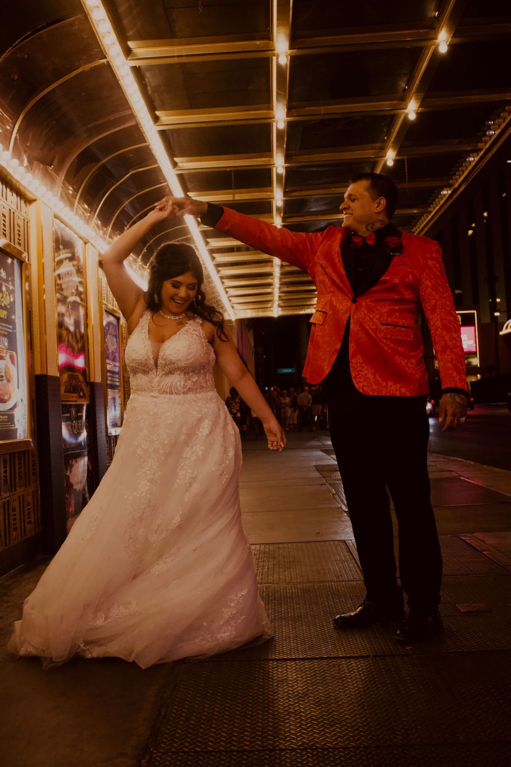 Bride in white gown twirls as groom in red sequin jacket holds her hand under glowing Las Vegas casino lights.