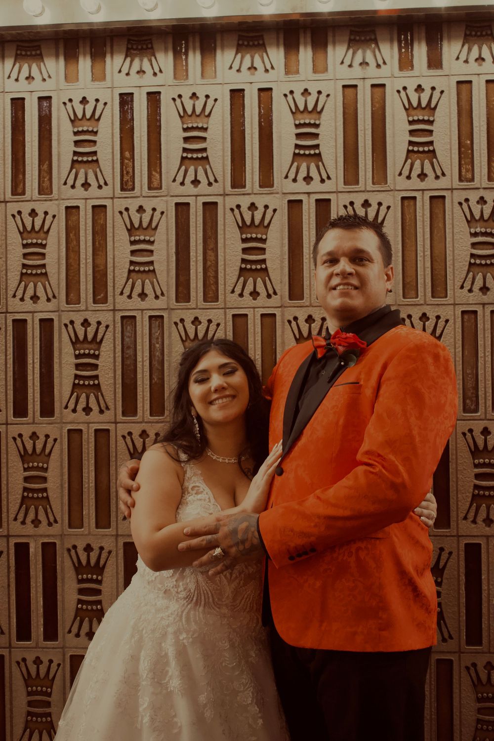 Bride in white gown and groom in orange blazer pose together in front of decorative crown-patterned wall.