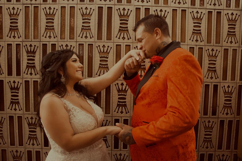 Groom in orange suit kissing bride's hand at wedding ceremony with decorative patterned backdrop.