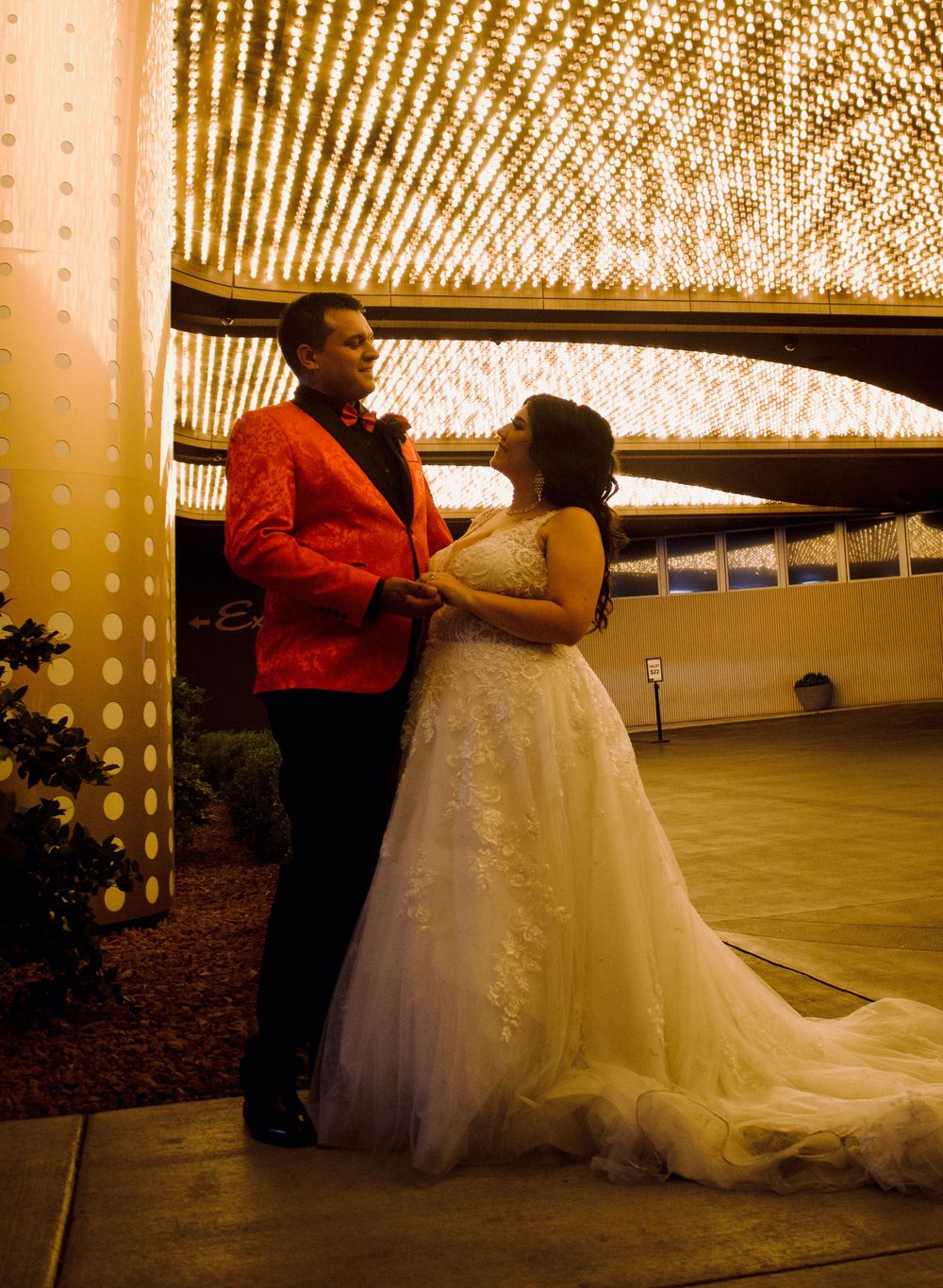 Bride in lace gown and groom in red blazer share romantic moment under glittering lights at Las Vegas wedding.