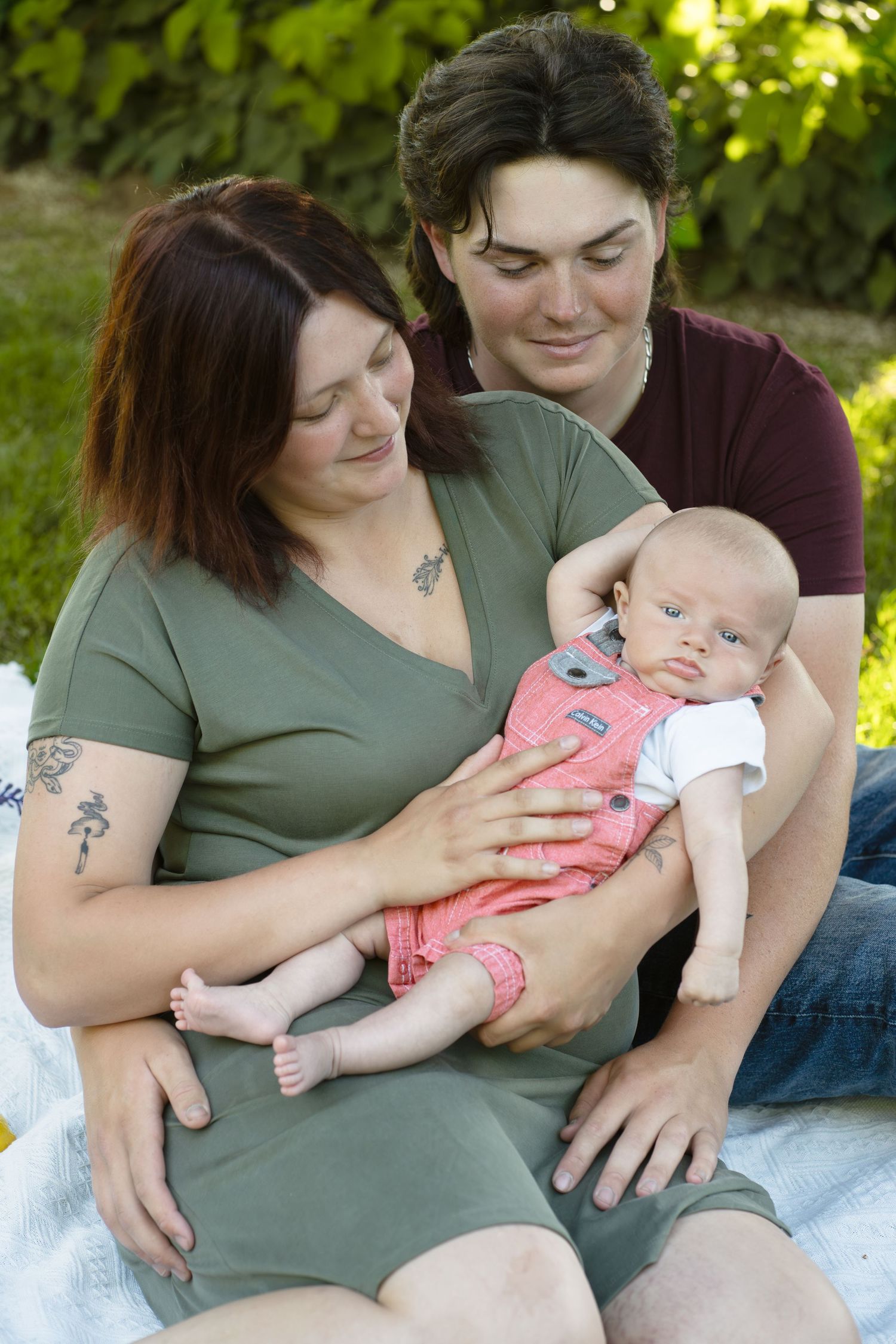 A family sitting on a blanket outdoors with their baby during a summer day.